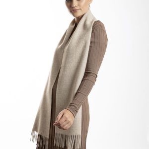 Woman wearing an oatmeal alpaca scarf from Tukuy with fringed edges, styled over a beige dress in a white background