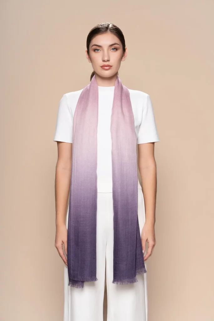 Woman wearing a gradient pink-to-purple baby alpaca wool scarf from Tukuy, styled over a white outfit and posed against a soft beige background.