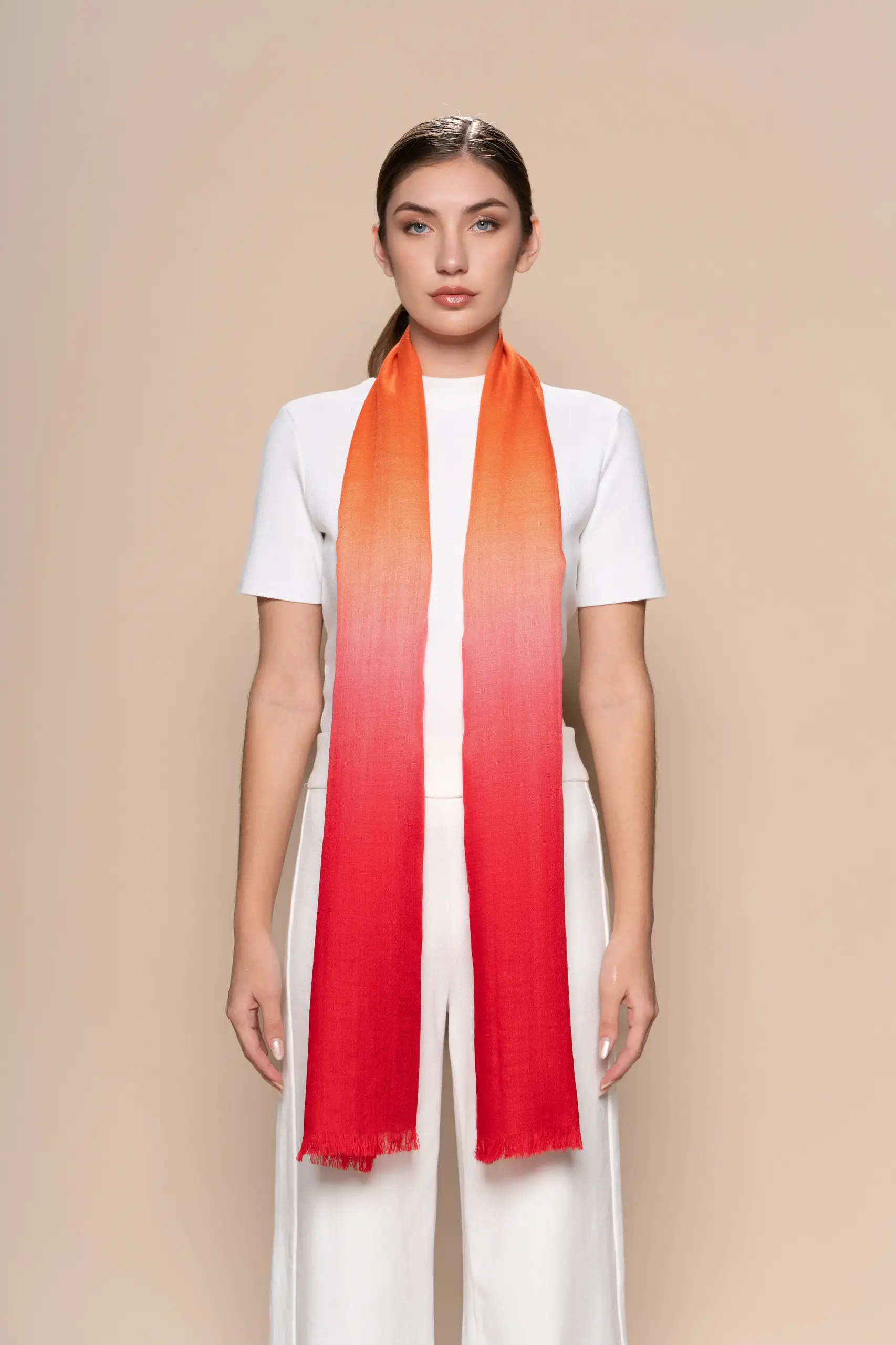Woman wearing a gradient orange to red baby alpaca shawl from Tukuy, styled over an all-white outfit against a neutral beige background.