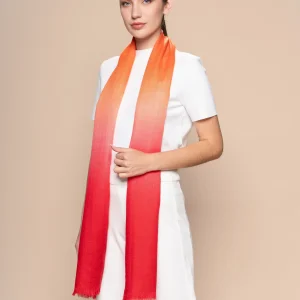 Woman wearing a gradient orange to red baby alpaca shawl from Tukuy, styled over an all-white outfit against a neutral beige background.