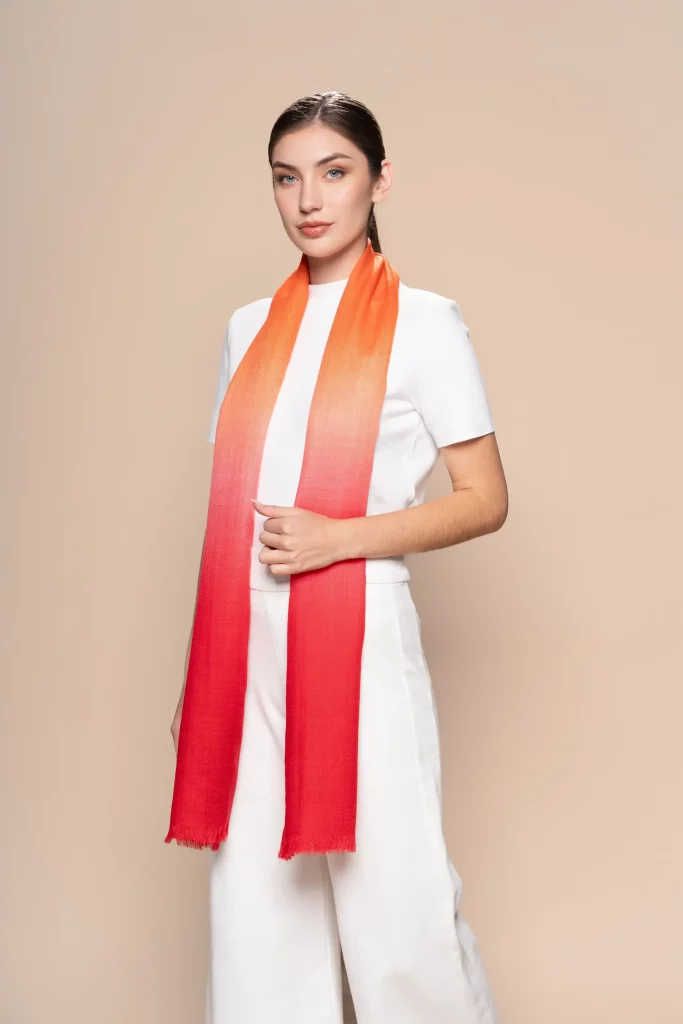 Woman wearing a gradient orange to red baby alpaca shawl from Tukuy, styled over an all-white outfit against a neutral beige background.