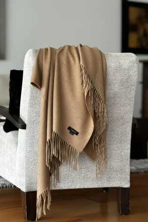 Camel-colored baby alpaca wool blanket from Tukuy draped over a textured grey armchair in a cozy and elegant living room setting.