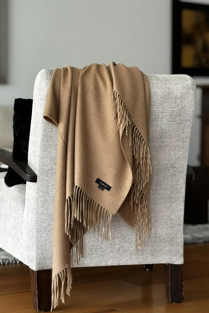 Manta de llana de baby alpaca de color camel de Tukuy sobre una butaca grisa texturitzada en un ambient de sala d'estar acollidor i elegant.
