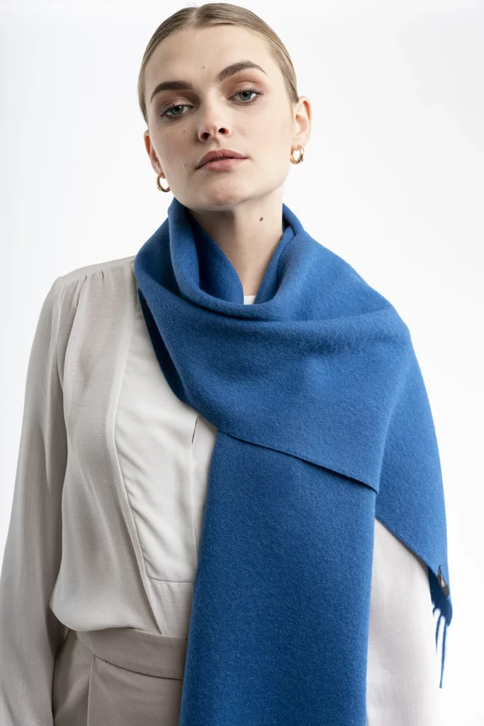 Woman wearing a blue alpaca scarf from Tukuy with fringed edges, styled over a white outfit against a neutral background.