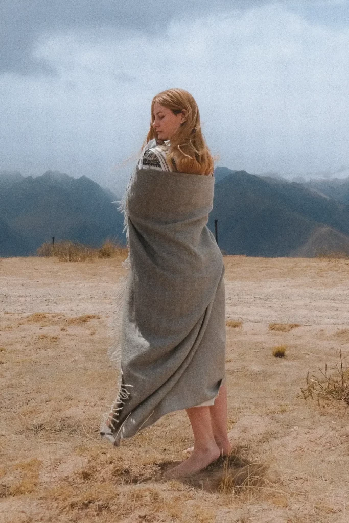Mujer vestida con una manta gris de lana de baby alpaca de Tukuy, de pie y descalza en la cordillera de los Andes, con los picos nevados al fondo.