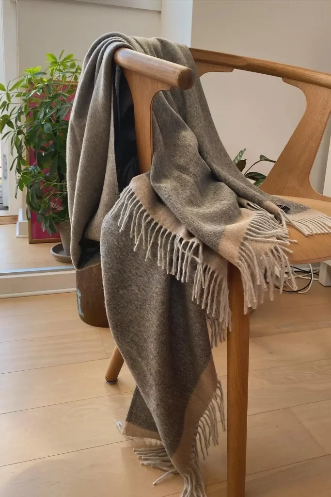 Suave manta de alpaca gris y beige de Tukuy con bordes y flecos beige, tapizada sobre una silla de madera en una acogedora habitación iluminada por el sol y con plantas de interior.
