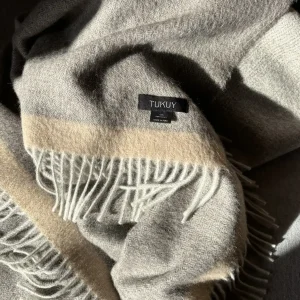 Soft grey baby alpaca blanket from Tukuy with beige stripe and fringed edges, folded in natural light to highlight its texture and warmth.