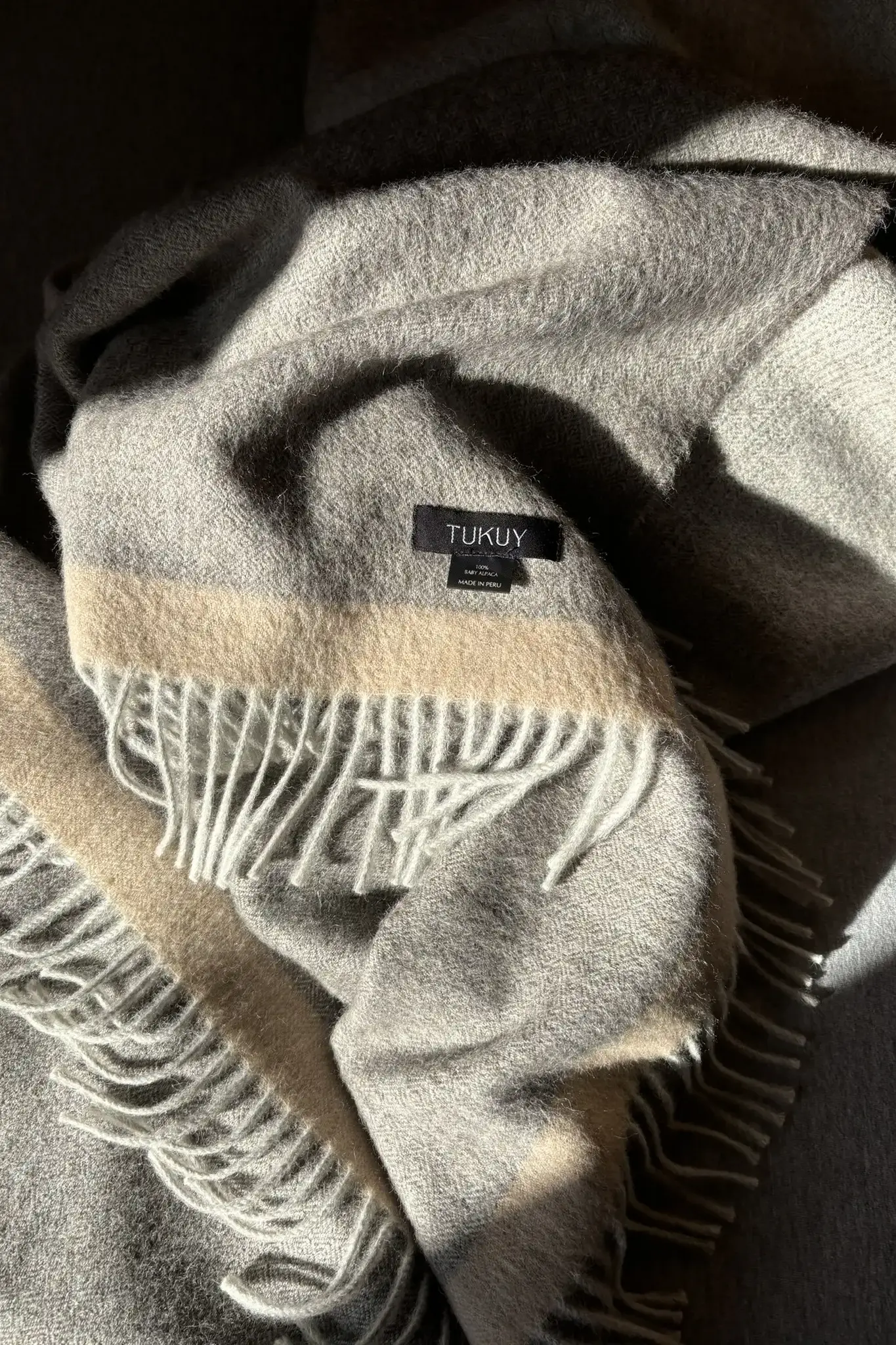 Soft grey baby alpaca blanket from Tukuy with beige stripe and fringed edges, folded in natural light to highlight its texture and warmth.