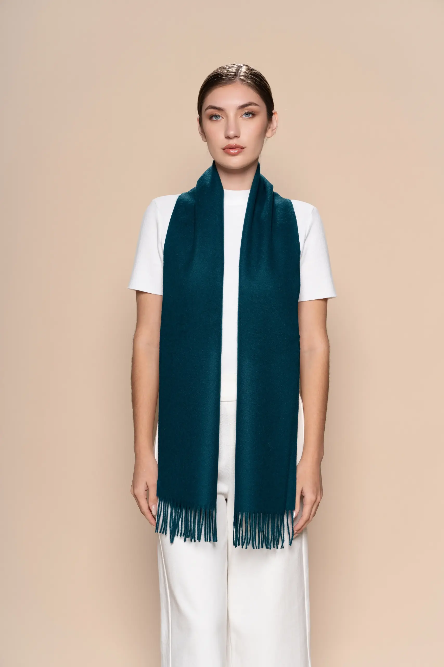 Woman wearing a deep teal baby alpaca wool scarf from Tukuy with fringed edges, styled over a white outfit against a neutral beige background.