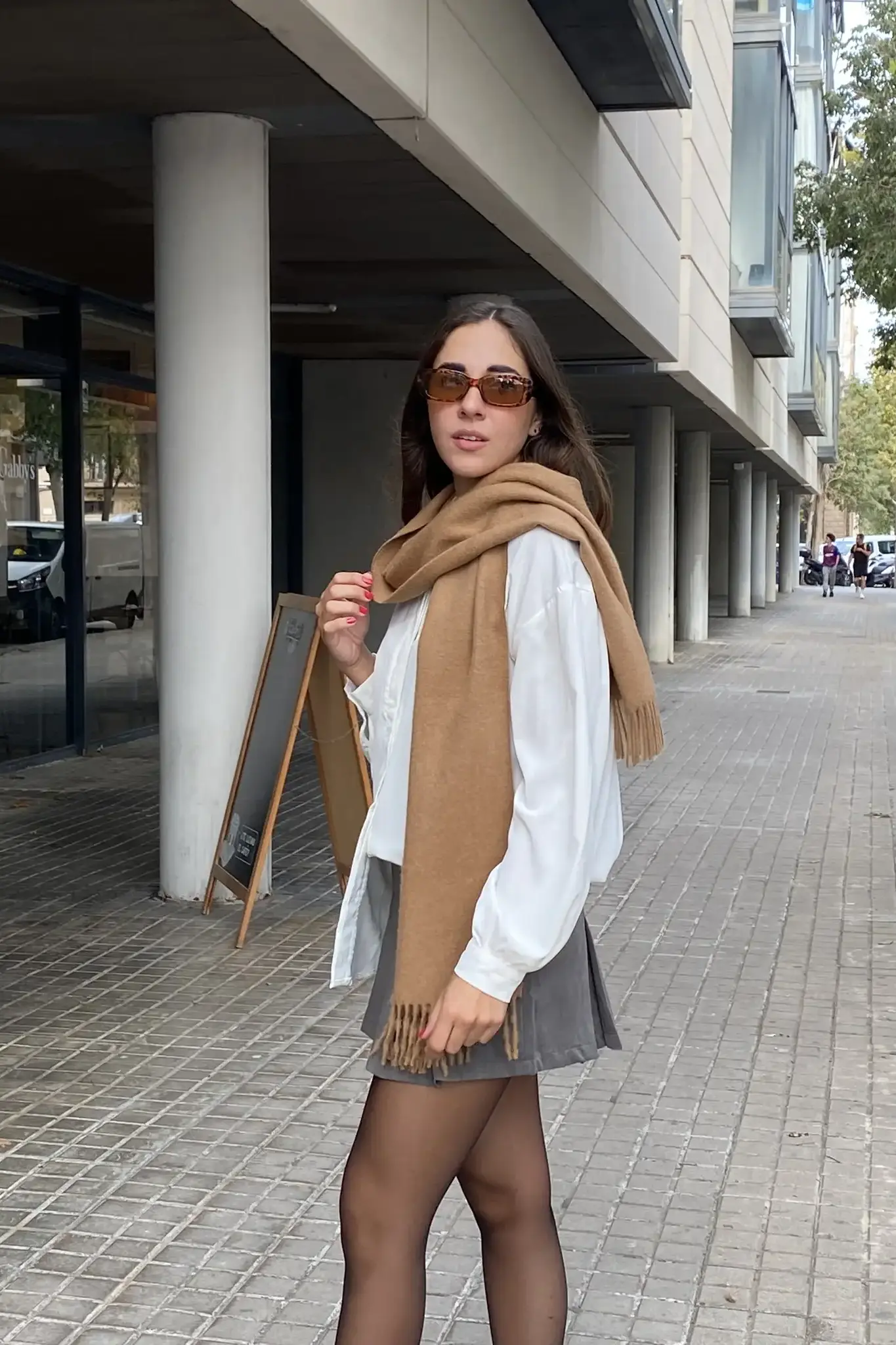 Woman wearing a camel-colored baby alpaca scarf from Tukuy with fringed ends, styled over a white blouse and sunglasses in an urban setting.