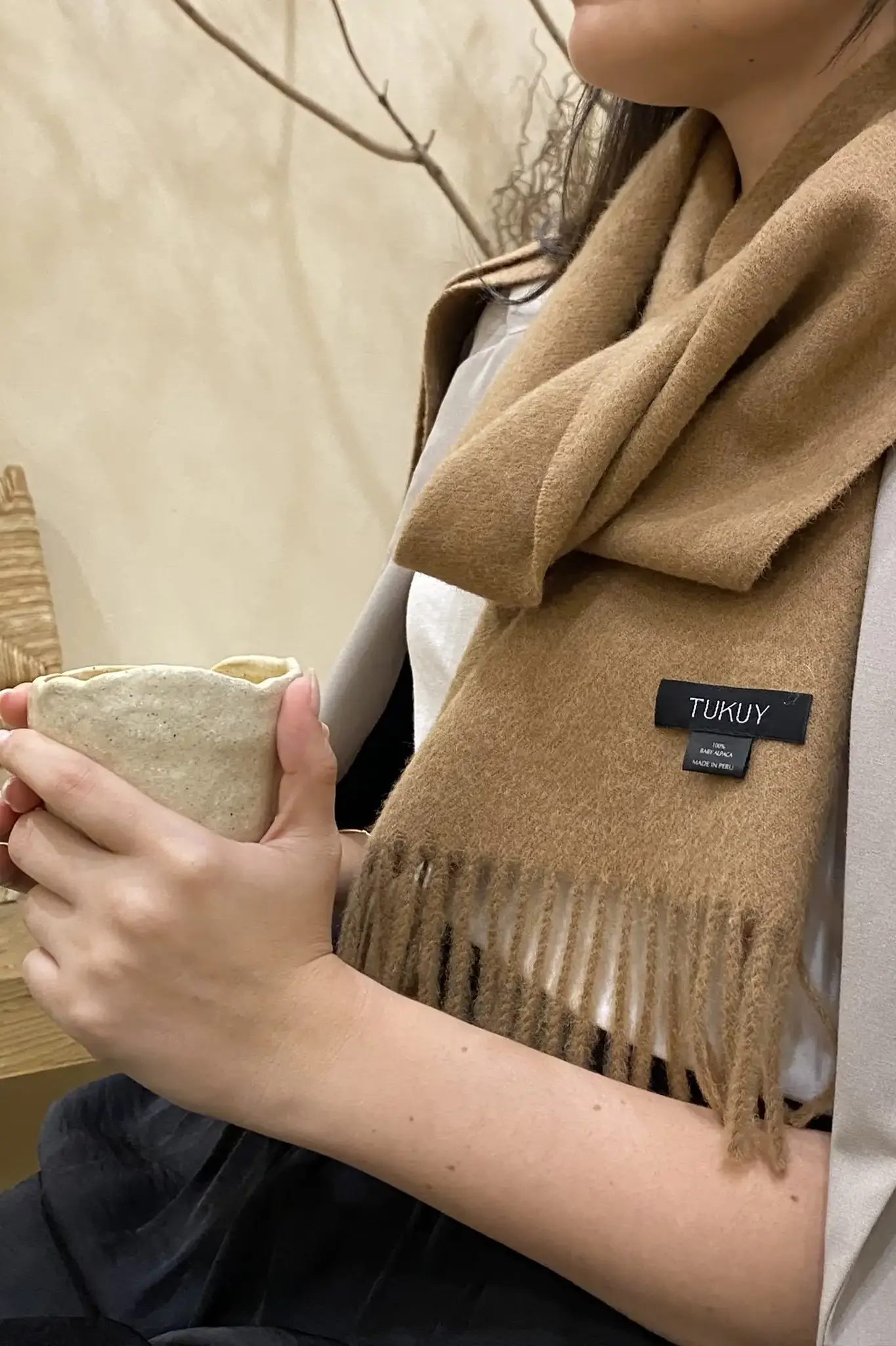 Woman holding a ceramic cup while wearing a camel-colored alpaca scarf from Tukuy, with fringed edges and a cozy, minimalist style.