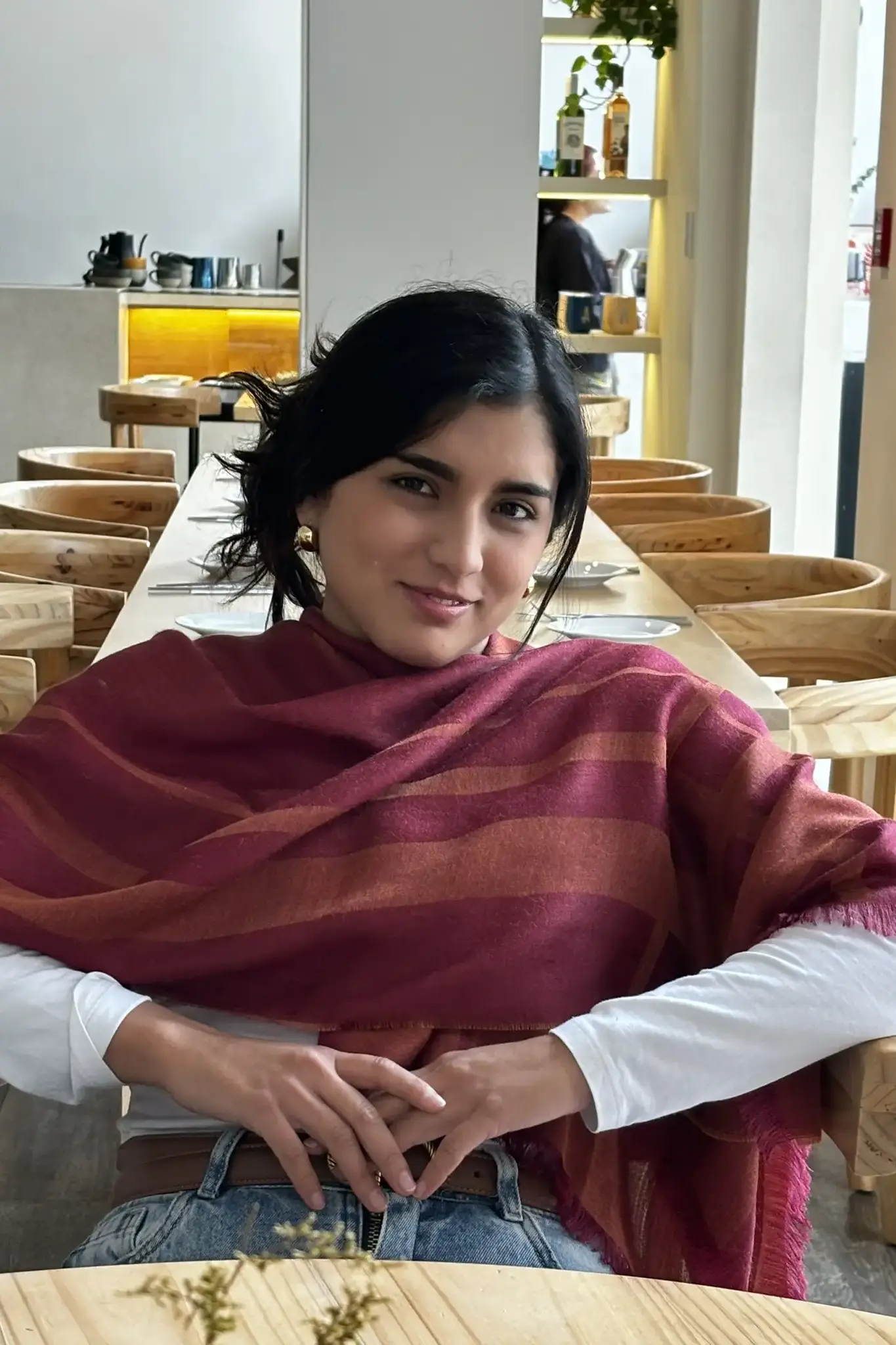 Woman sitting at a wooden table wearing a striped burgundy baby alpaca wool shawl from Tukuy, styled casually over a white top and jeans.