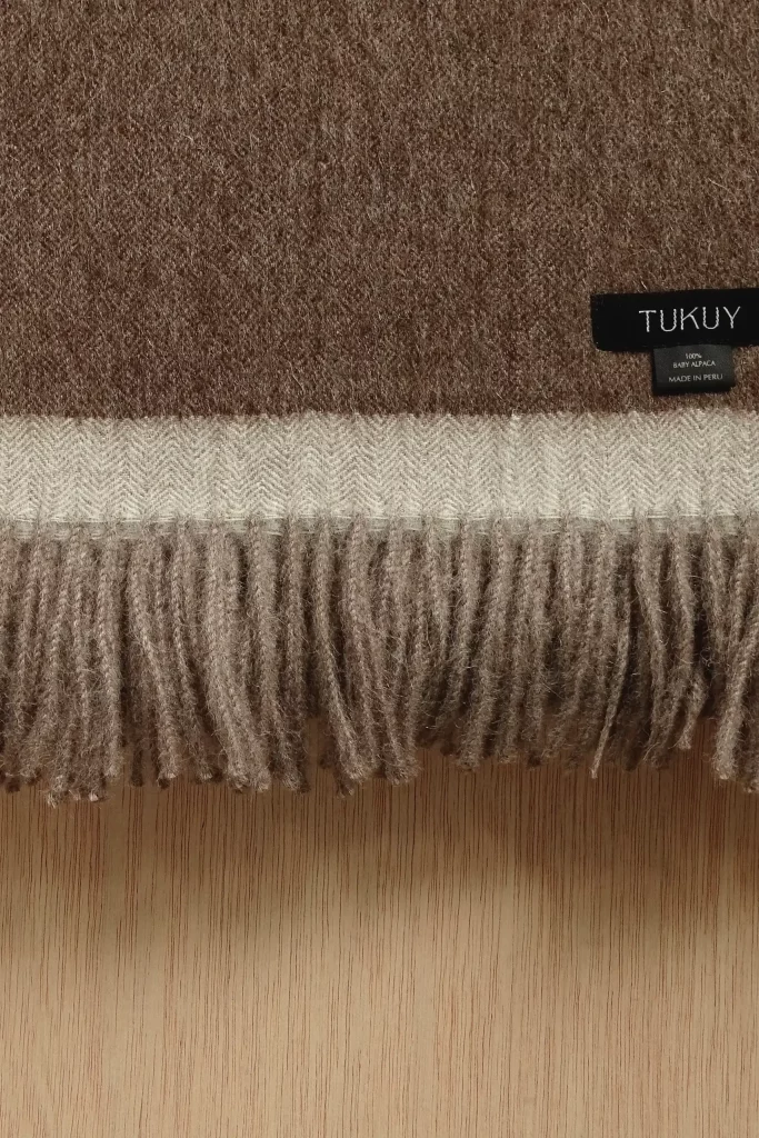 Primer plano de una manta de lana de baby alpaca marrón de Tukuy con detalle de flecos crema y etiqueta tejida, colocada sobre una superficie de madera