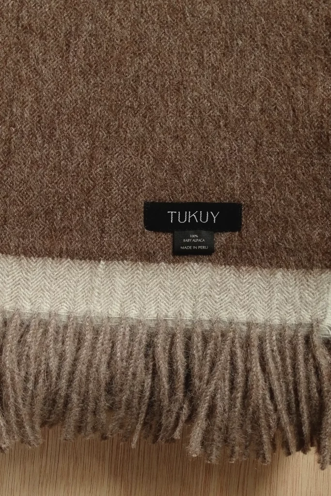 Primer plano de una manta baby alpaca marrón y beige de Tukuy con detalle de flecos crema y etiqueta tejida, colocada sobre una superficie de madera.
