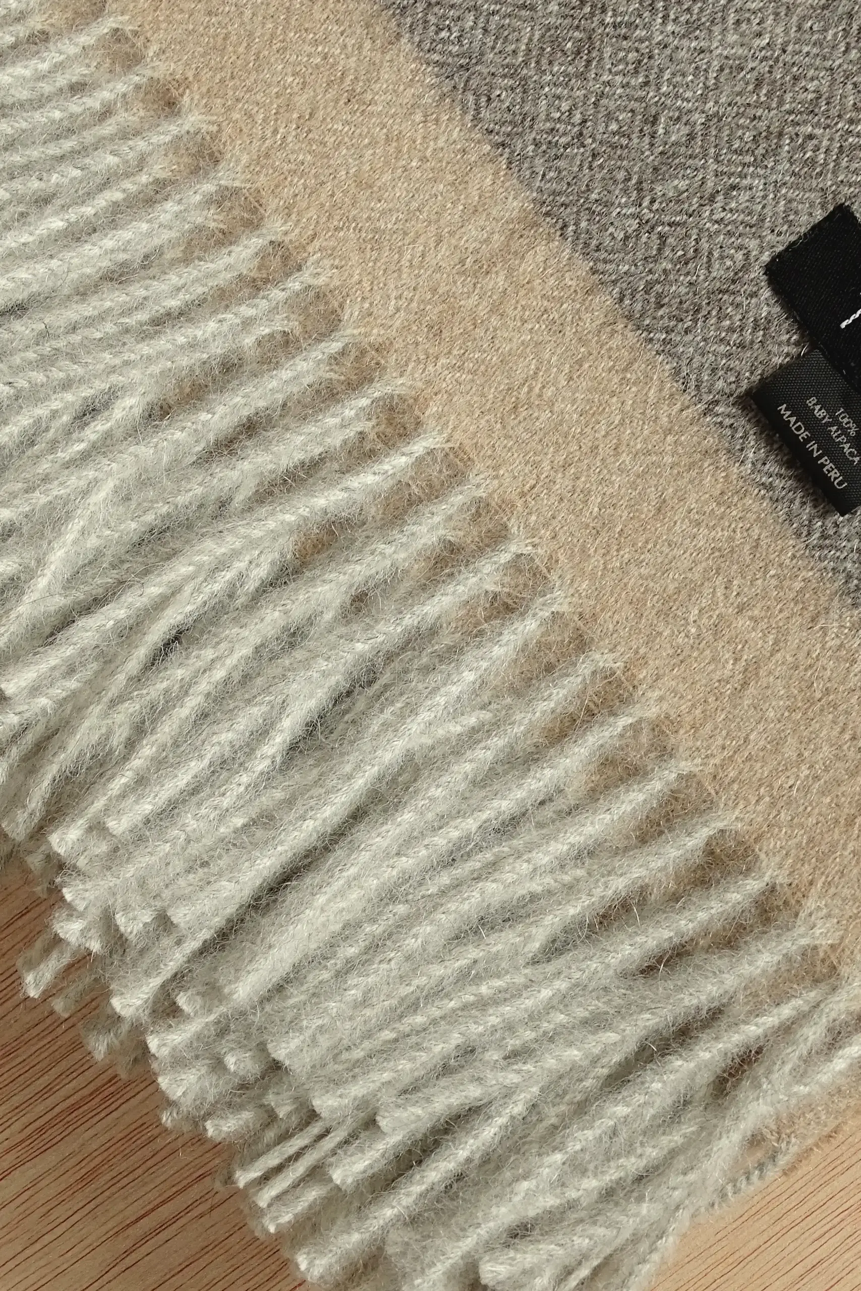 Close-up of a gray baby alpaca wool blanket from Tukuy with beige fringe detail and woven label, placed on a wooden surface.