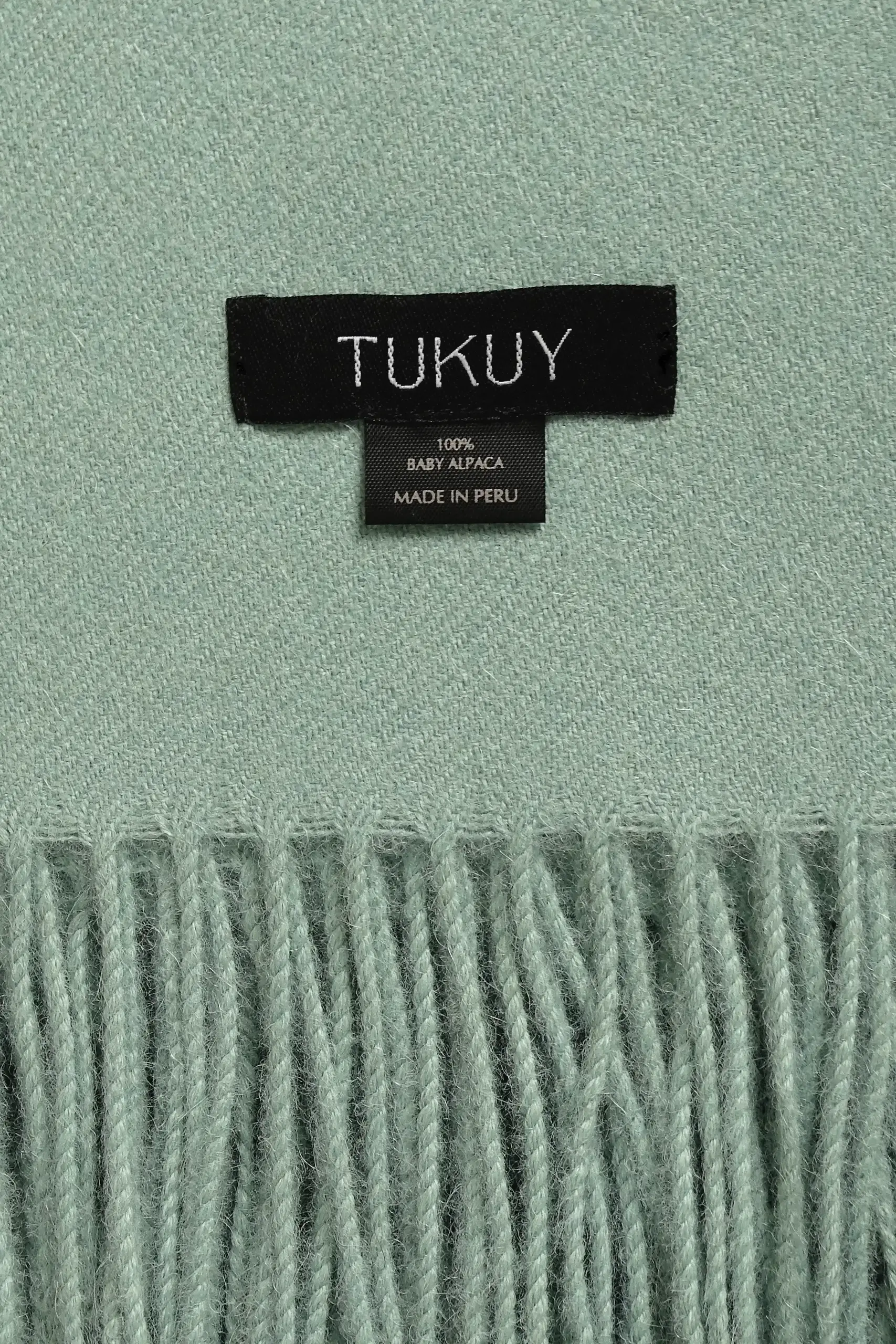 Close-up and detailed picture of a soft green alpaca blanket from Tukuy with fringed edges and a black woven label 100% baby alpaca made in Peru, placed on a wooden surface.