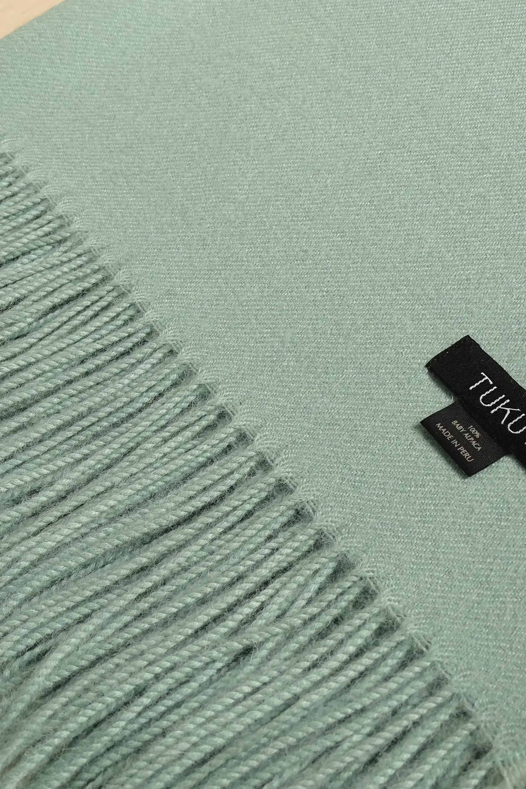 Close-up of a soft green alpaca blanket from Tukuy with fringed edges and a black woven label, placed on a wooden surface.