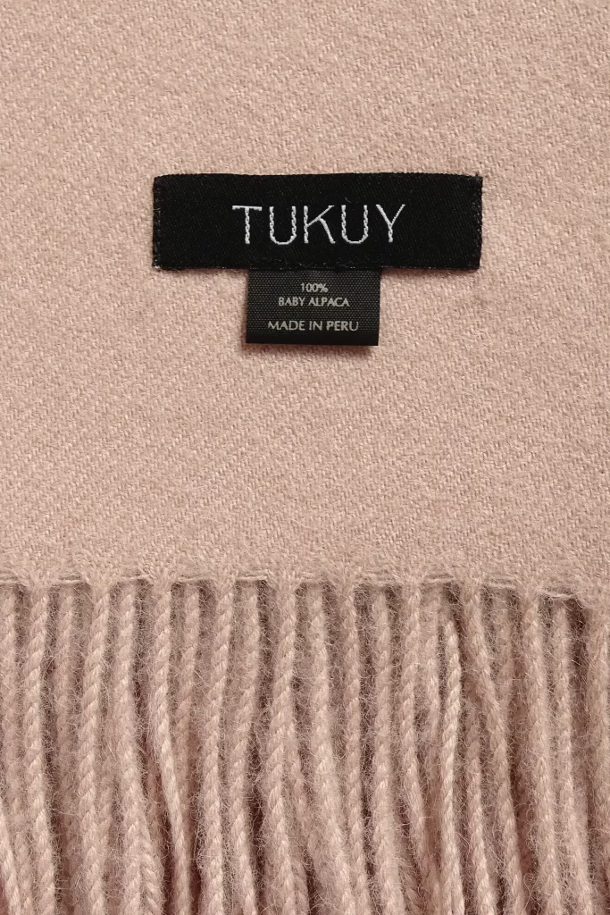 Primer plano de una manta de alpaca rosa de Tukuy con detalle de flecos y etiqueta tejida, colocada sobre una superficie de madera.