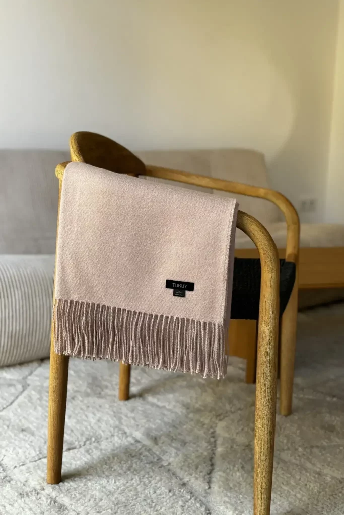 Manta de lana rosa claro baby alpaca de Tukuy sobre una silla de madera en un salón moderno con un sofá beige y una mesa de centro redonda de madera.