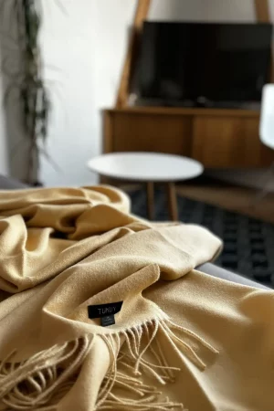 Yellow baby alpaca wool blanket from Tukuy draped over a sofa in a cozy living room, with wall art and a potted plant in the background.