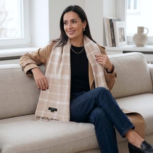 Woman styling a beige check alpaca scarf with a camel jacket, black top, and blue jeans, demonstrating the versatility of the scarf's heritage-inspired pattern with casual and sophisticated outfits.