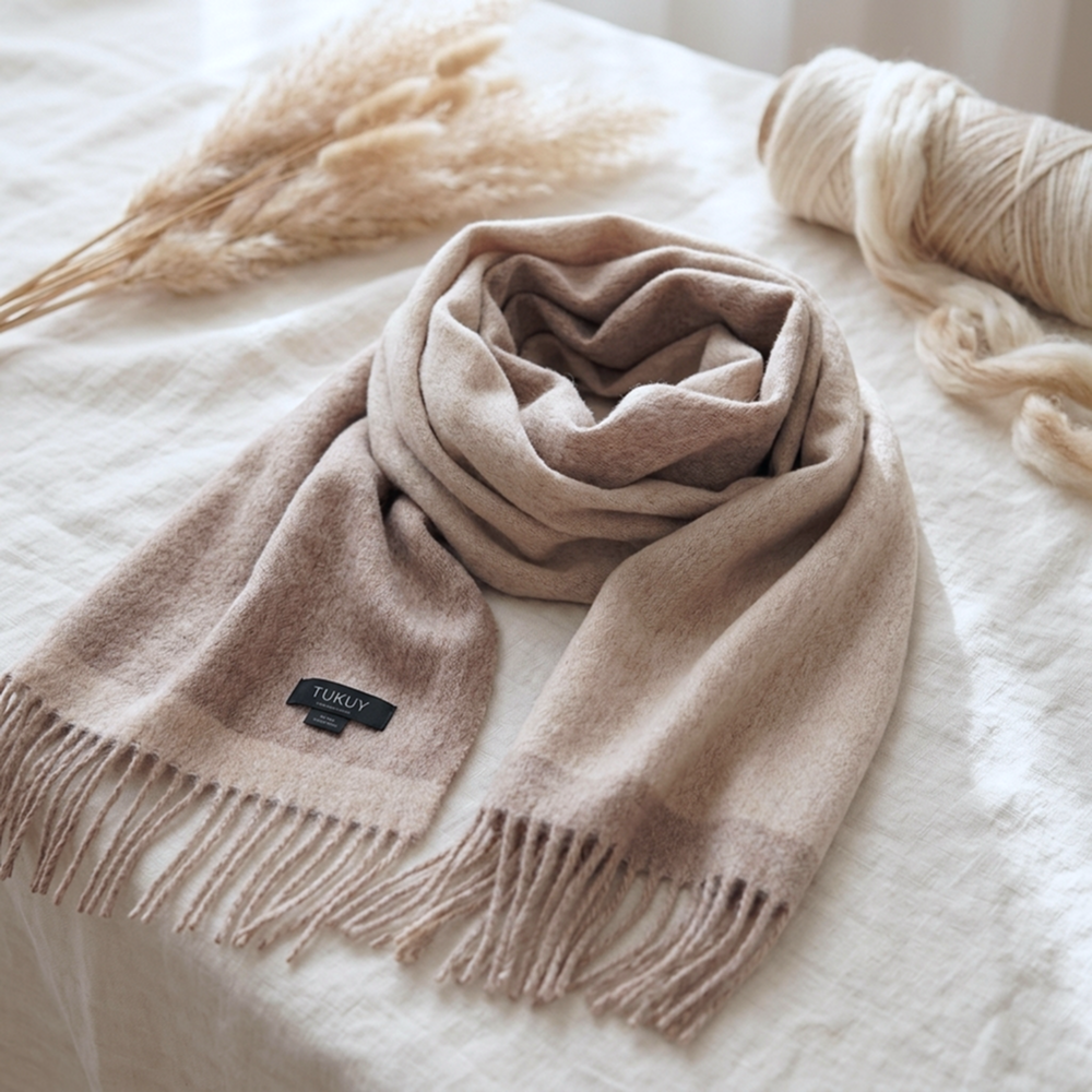 Ein beidseitig tragbarer Schal aus Alpakawolle in Beige und Taupe, präsentiert auf einem neutralen Leinenhintergrund, der seine natürlichen Farben, seine luxuriöse Weichheit und die von Hand gearbeiteten Fransen besonders zur Geltung bringt.